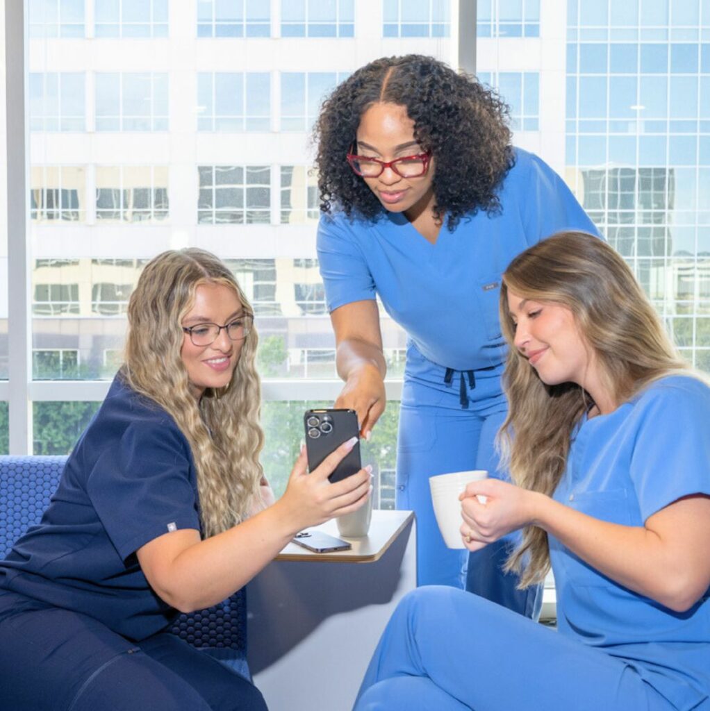 Three healthcare professionals in scrubs, gathered around a small table, looking at a smartphone. One woman is pointing at the phone while the other two smile, with one holding a coffee mug. They are in a bright room with large windows and a cityscape in the background