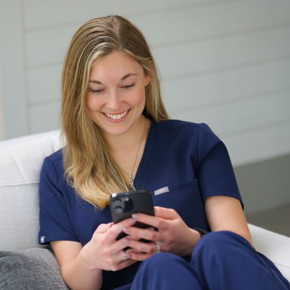 A healthcare professional in blue scrubs is seated on a grey couch, engrossed in a smartphone. The individual’s face is intentionally blurred for privacy