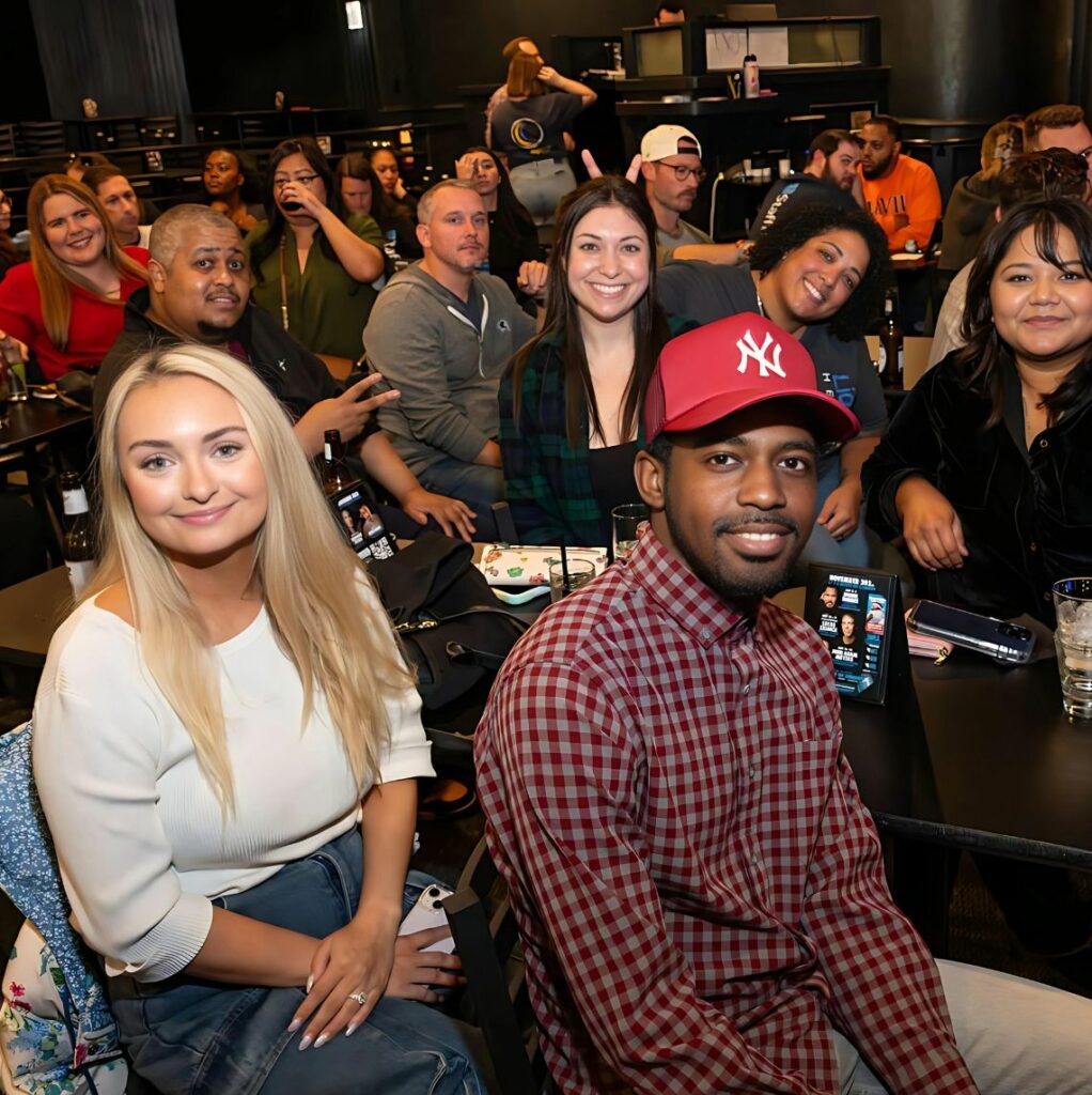 A group of people sitting together in a casual setting, smiling and looking at the camera. The group includes a mix of men and women, with one man wearing a red New York Yankees cap in the foreground. They appear to be at an event or gathering with drinks and menus on the tables