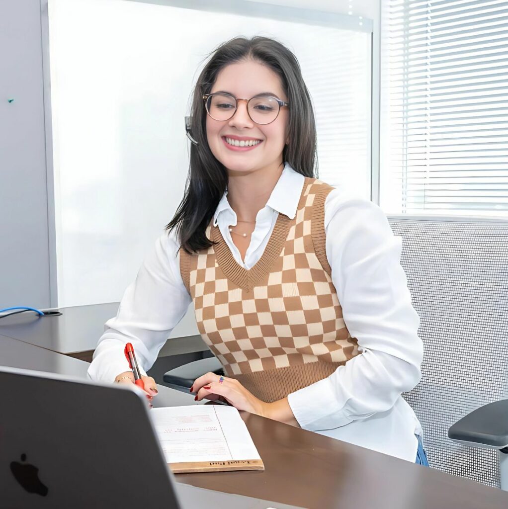 Smiling woman wearing glasses and a checkered sweater vest over a white shirt, sitting at a desk with a laptop and holding a pen, looking at documents